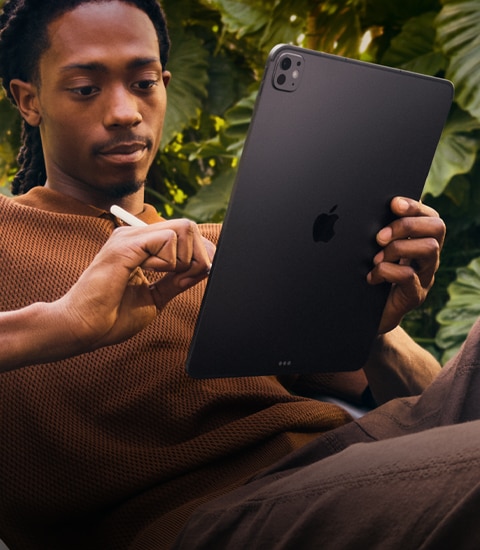 A person outside by foliage holding their iPad Pro in one hand and Apple Pencil Pro in the other
